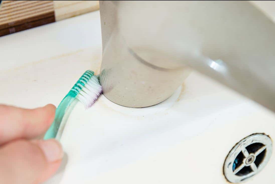 person using blue toothbrush to clean mould around bathroom sink faucet
