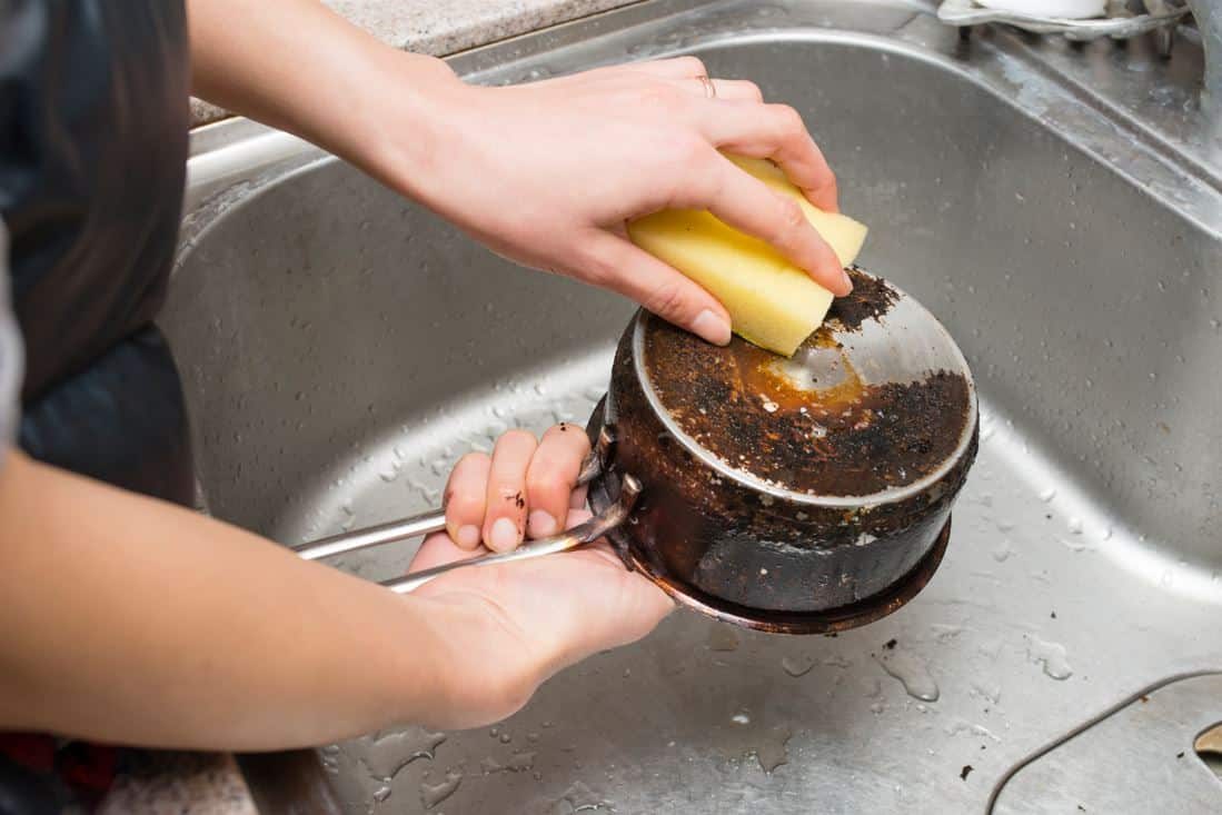 woman scraping the bottom of an dirty aluminium pot with a yellow sponge