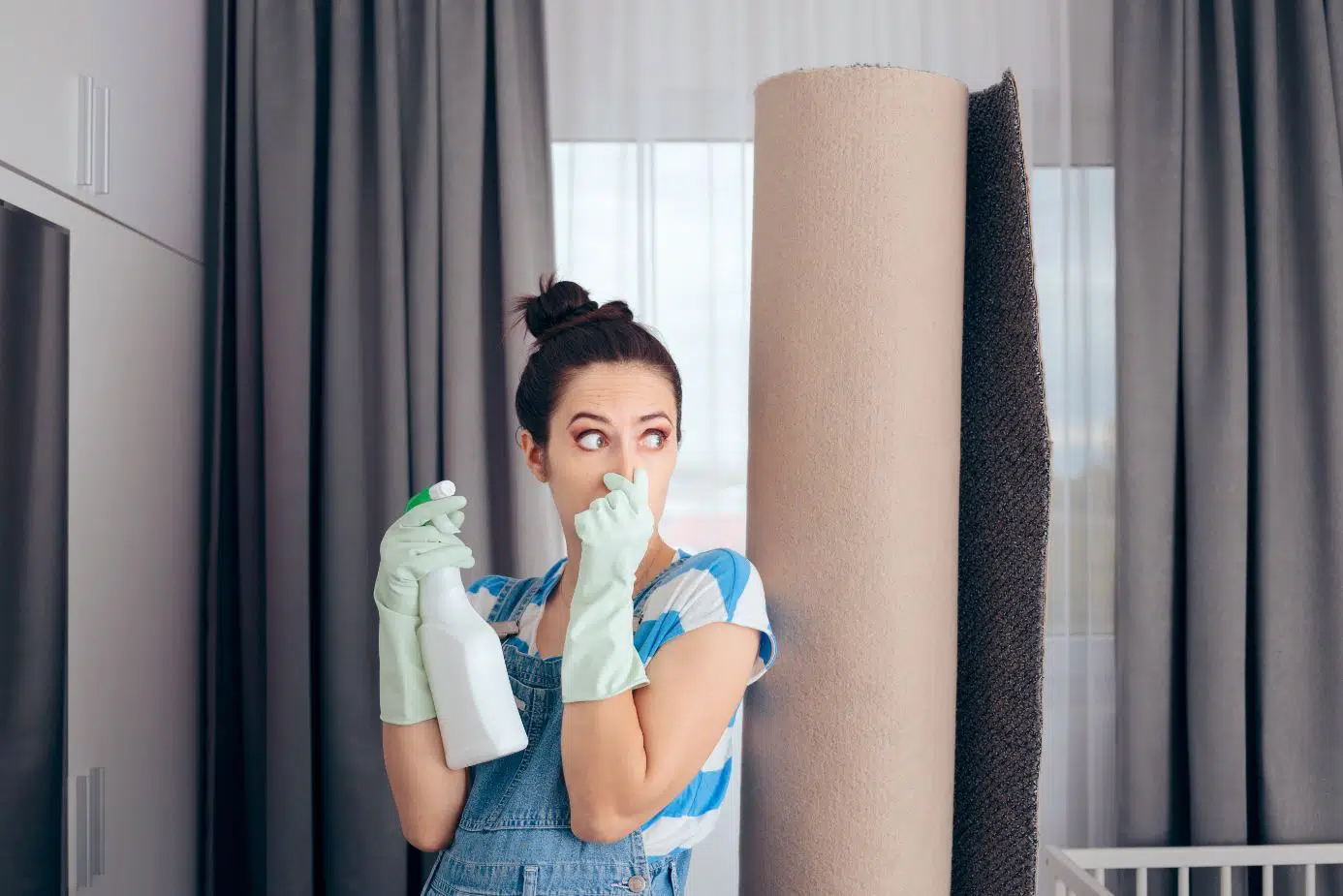 woman wearing blue overalls standing next smelly carpet holding her nose