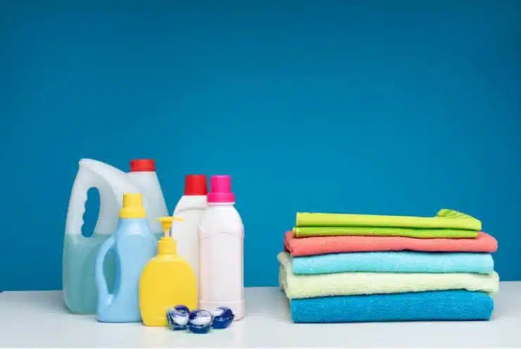five assorted coloured towels folded on top of each other next to six bottles of laundry detergent