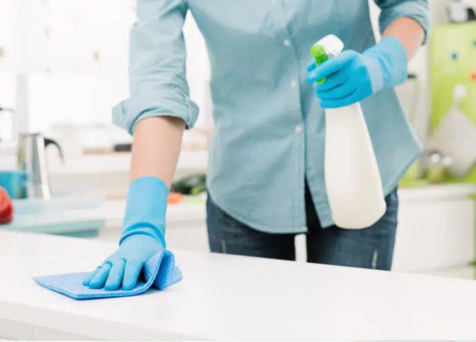 woman in blue collared shirt and jeans wiping down a white benchtop with a blue sponge