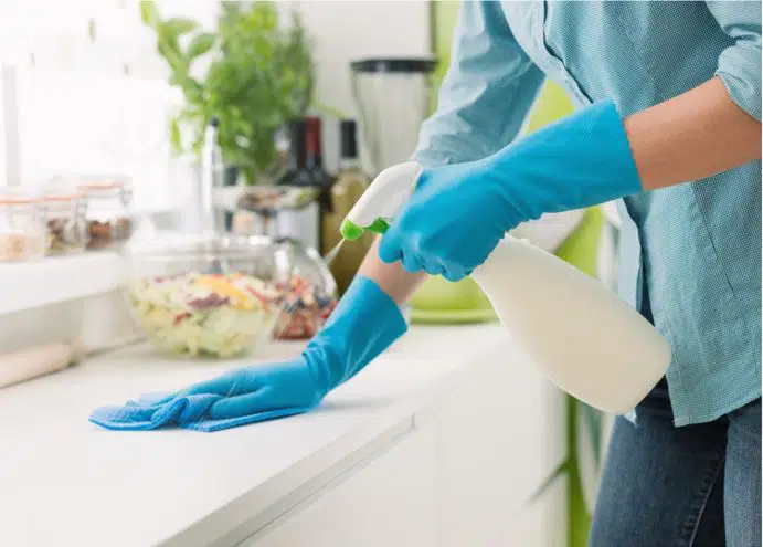 woman wearing blue gloves spraying down white kitchen benchtop with disinfectant