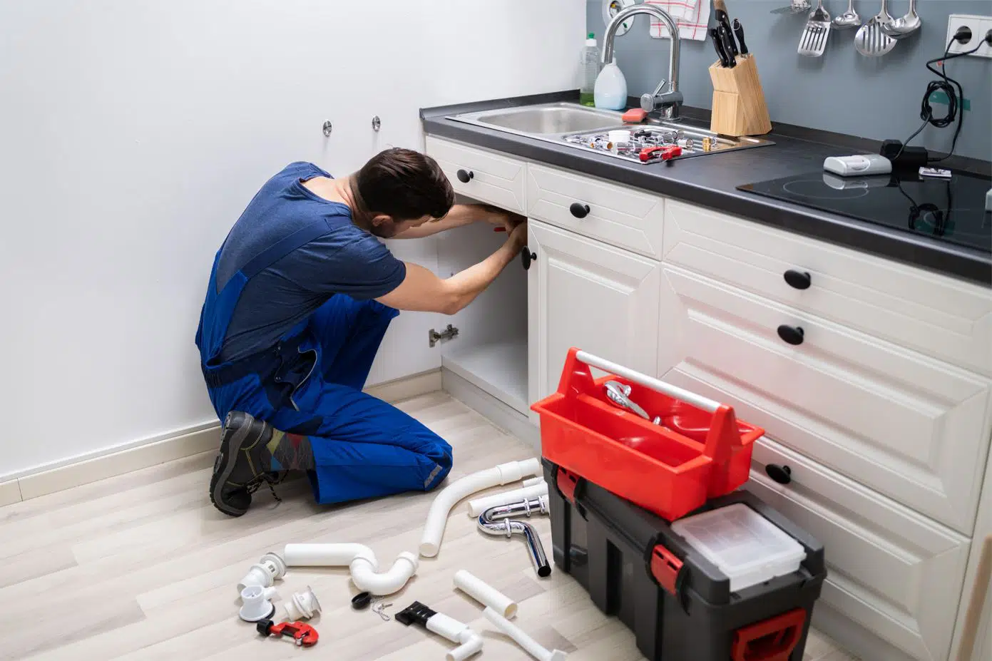plumber wearing blue overalls replacing kitchen sink drain pipe with tools next to him
