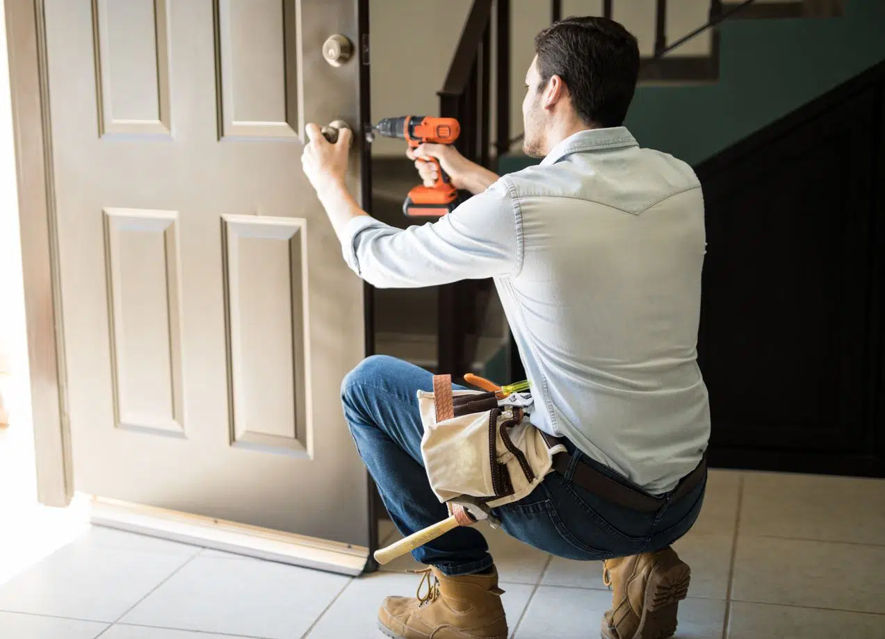 young black haired handyman with tool belt fixing a door mechanism with an orange drill