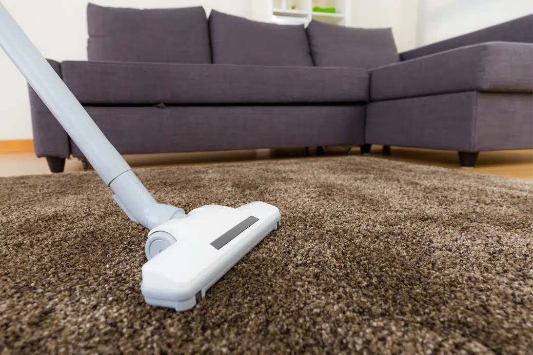 close up of white vacuum cleaning large brown shag rug with a 5 seater grey couch in the background