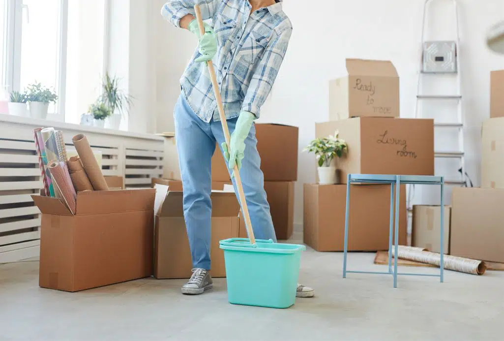 woman wearing blue shirt and blue pants holding a mop and bucket in an empty living room with moving boxes in the background