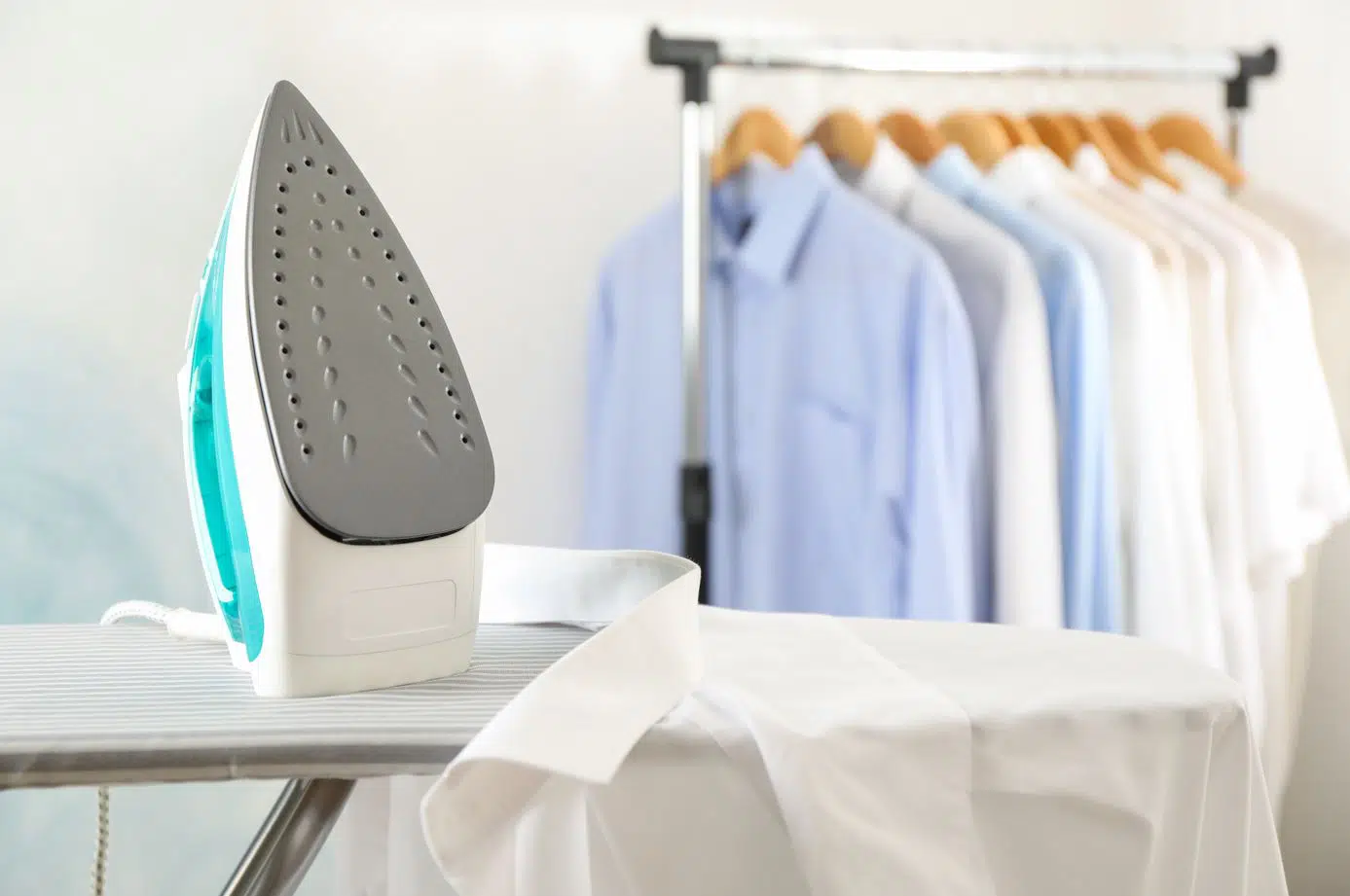 blue and white iron face up on a grey ironing board next to a white button up shirt with button up shirts in the background on a hanger