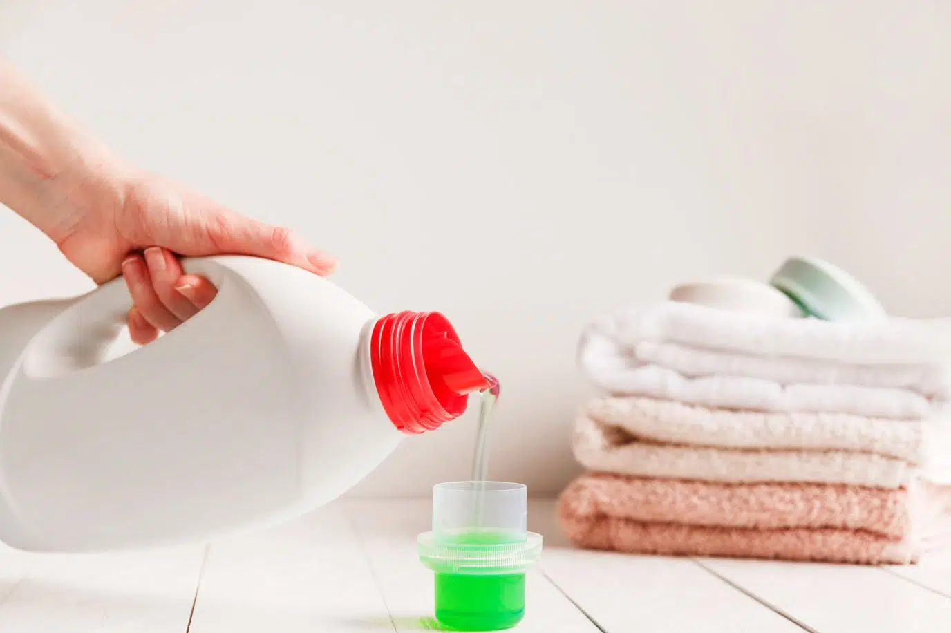 woman pouring green laundry detergent into laundry detergent cap with 3 folded towels in the background