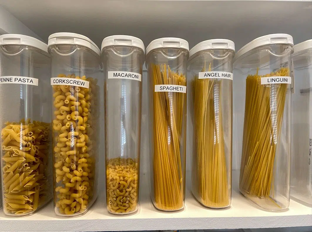 6 assorted plastic containers in an all white pantry containing different kinds of pasta