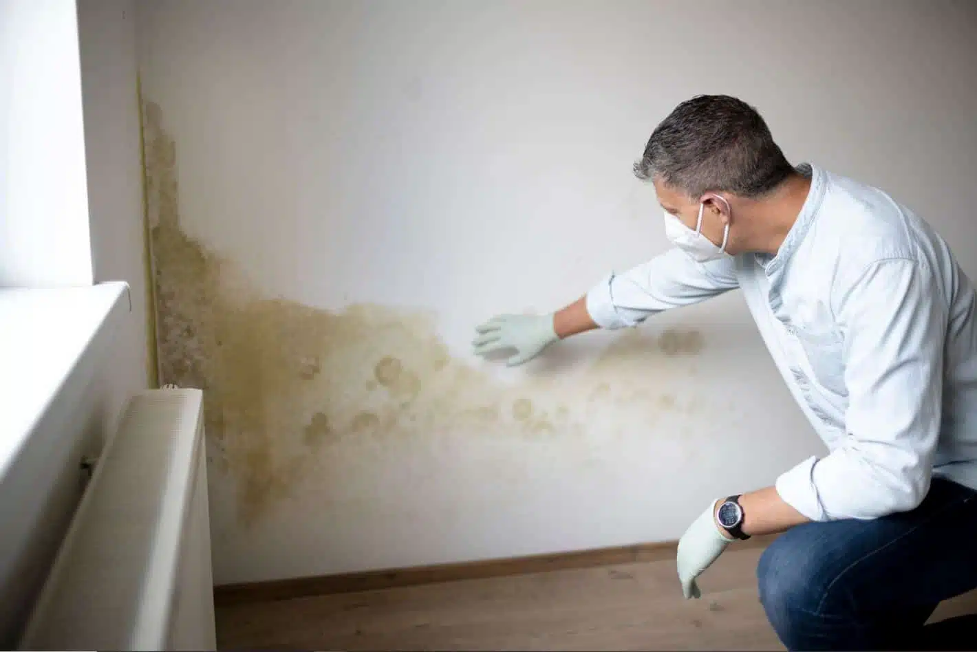 man wearing mask and protective gear inspecting mould on a white wall in an empty apartment