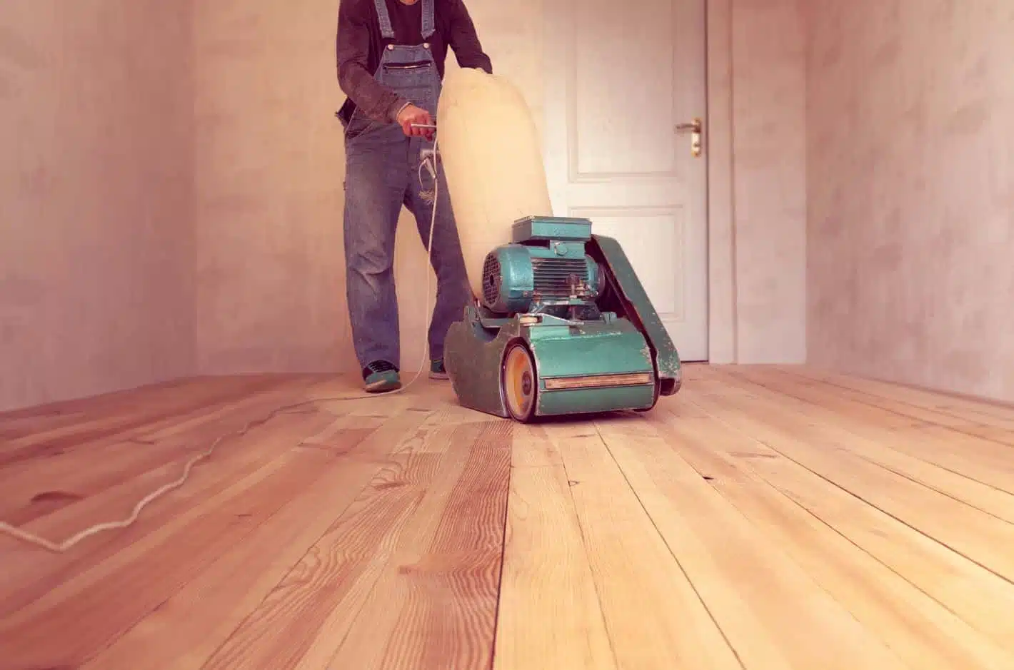man wearing overalls pushing an old green floor sander over a hardwood floor in an empty room