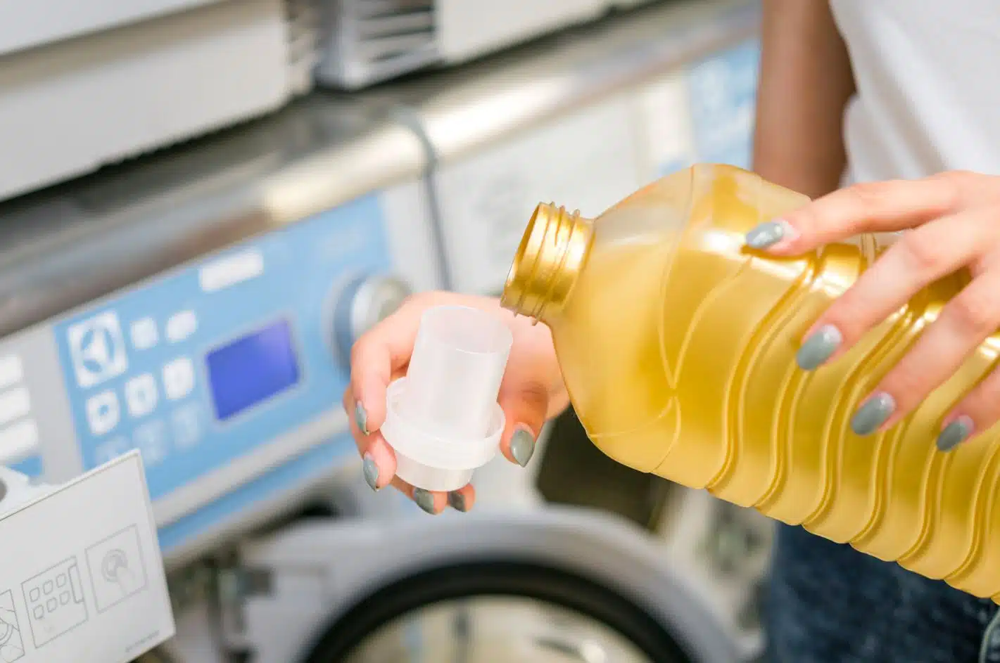 woman with blue nail polish holding gold laundry detergent container and pouring the liquid into the cap