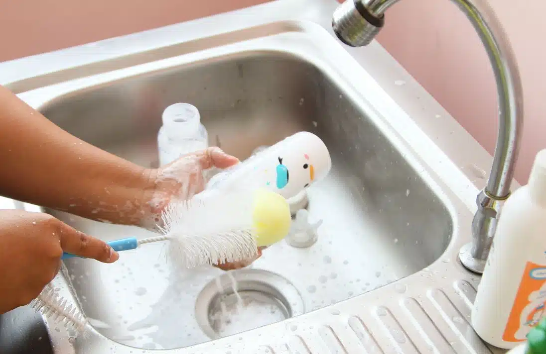 woman cleaning white babies bottle in a stainless steel sink