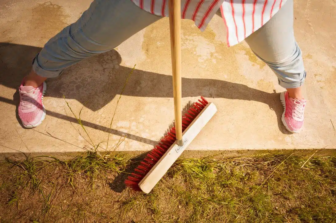 woman with pink shoes using a red bristled brush to clean the outside patio of her home