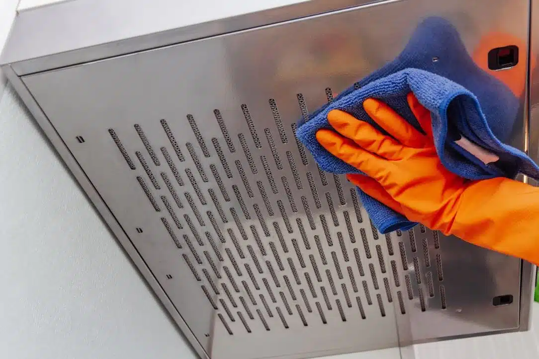 person wearing orange gloves using a blue microfibre cloth to clean rangehood and filter